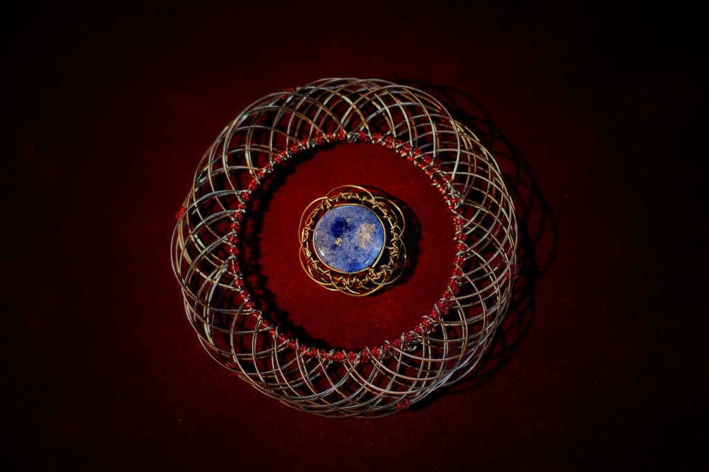 Un grand mandala en fils argentés reliés par des perles rouges entoure un plus petit avec une pierre bleue. Les deux bijoux sont posé sur un fond bordeaux.