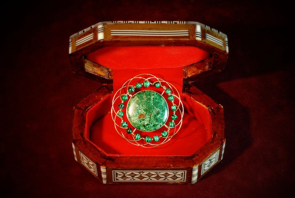 Mandala manufacturé par Jonas avec une pierre verte sertie en son centre. Le bijou repose dans un écrin nacré garni de velours rouge.
