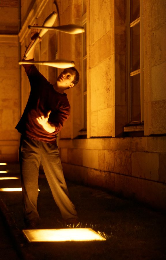 Martin fait de la jonglerie à 3 massues derrière l'Hôtel de ville de Caluire-et-Cuire. Il est éclairé par les projecteurs au sol.