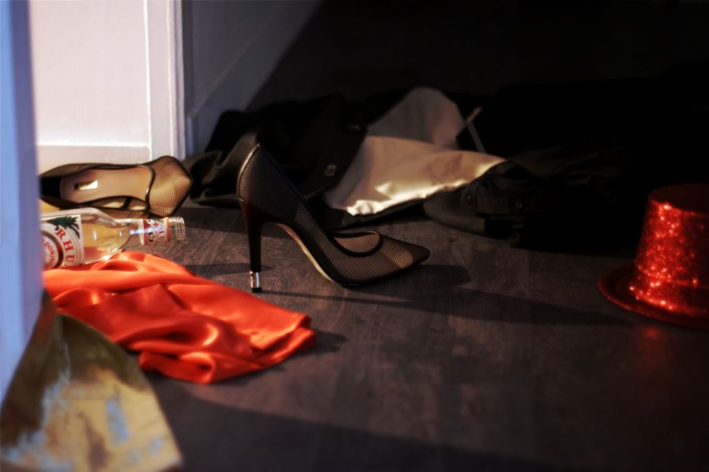 A l'entrée d'une chambre, des vêtements d'homme et de femme sont par terre dont une paire de talons aiguilles. Il y a aussi une bouteille d’alcool ouverte.