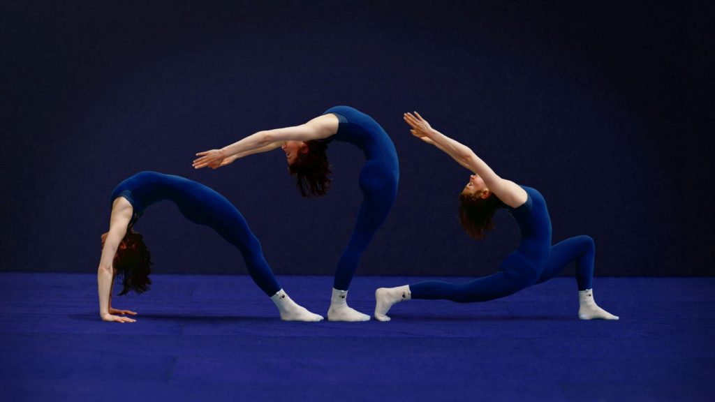 Gabrielle, en échauffement au gymnase d'Overground Circus utilise des positions de yoga. Trois positions sont visibles sur cette photo dans les tons bleus.