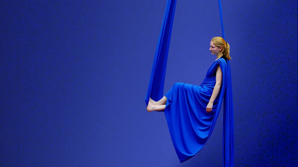 Vanille est assise en suspension dans un tissu aérien au gymnase d'Overground Circus. Le tissu bleu est replié en forme de robe. 