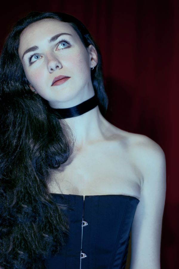 Portrait d'Iris en corset devant le rideau en satin bordeaux du boudoir. Elle porte un ruban de noir autour du cou qui fait ressortir la pâleur de sa peau.