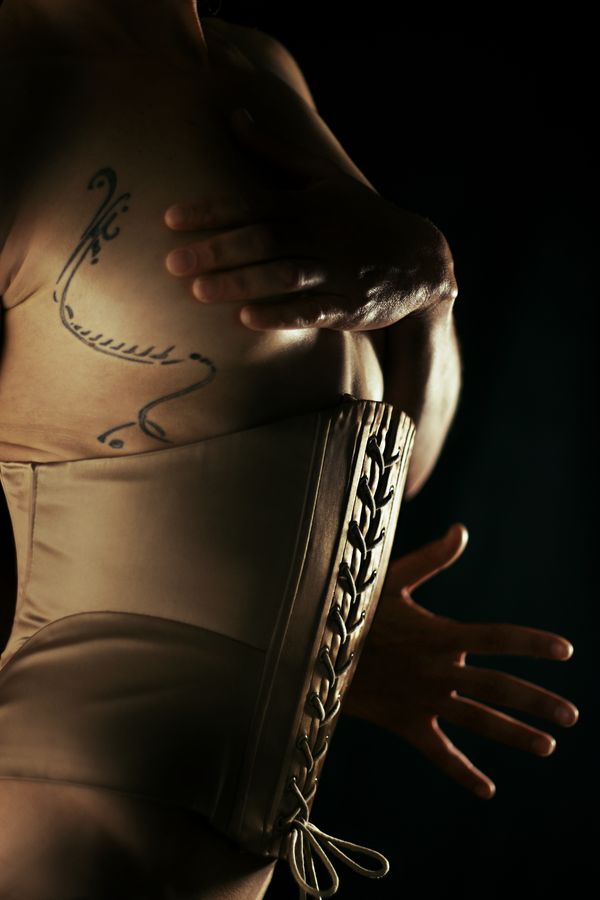 Céline, en corset laçé, cache sa poitrine pour ne dévoiler que le tatouage qui fait le tour de son sein. Son autre main dépasse derrière elle.